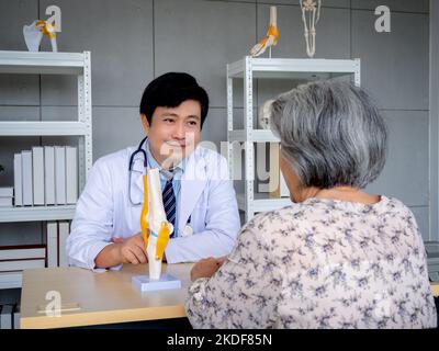 Homme asiatique souriant médecin orthopédique en costume blanc pointant vers le modèle anatomique de l'articulation du genou à expliquer pour la patiente senior en médecine de Banque D'Images