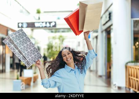 Bonne cashopper femme regardant l'appareil photo et souriant, hispanique femme avec la danse des cheveux bouclés et le saut avec plaisir, a acheté des cadeaux en vente, portrait de la bonne femme de shopping. Banque D'Images