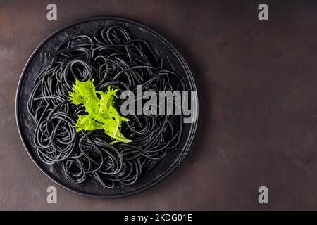 Spaghetti noirs sur fond sombre. Pâtes à l'encre de seiches noires bouillies avec laitue verte sur une assiette noire. Copier l'espace Banque D'Images