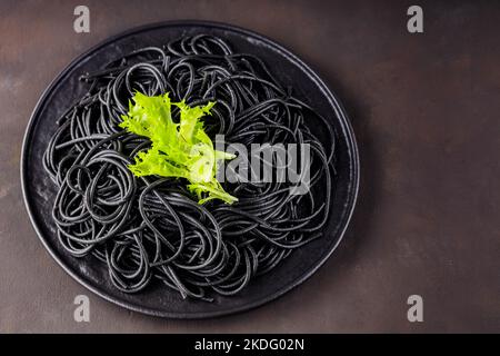 Spaghetti noirs sur fond sombre. Pâtes à l'encre de seiches noires bouillies avec laitue verte sur une assiette noire. Copier l'espace Banque D'Images