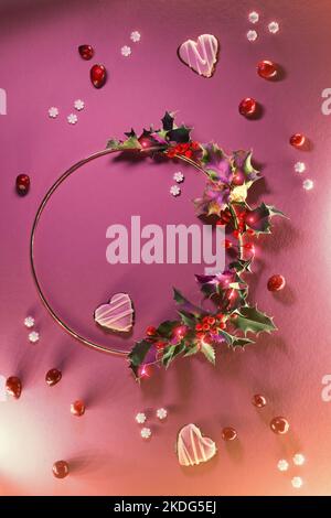 Plat de Noël en magenta et rouge. Couronne de Noël avec feuilles de houx et baies sur anneau métallique. Vue de dessus avec branches de sapin naturel, biscuits de coeur et cra Banque D'Images