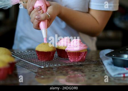 femme en chemise blanche étalant la crème de beurre rose givrant sur trois cupcakes, arrière-plan flou Banque D'Images