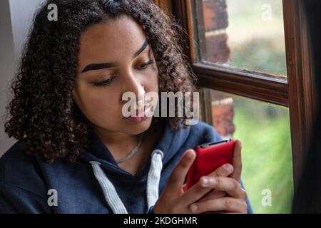 Belle triste biracial Africain American girl adolescente jeune femme triste dépressive ou réfléchie regardant hors d'une fenêtre en utilisant un téléphone cellulaire ou intelligent Banque D'Images