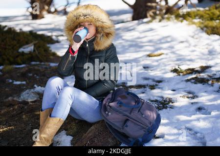 Femme buvant quelque chose de chaud d'une bouteille de thermos en métal assis sur un rocher dans les montagnes enneigées. Banque D'Images