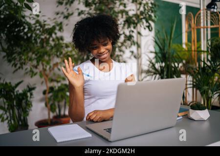 Une étudiante afro-américaine amicale se fait un bonjour à la webcam de l'ordinateur portable, parlant lors d'une vidéoconférence avec un professeur dans une maison lumineuse. Banque D'Images