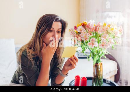 Une jeune femme surprise lit une carte laissée dans un bouquet de fleurs par un petit ami à la maison. Cadeau avec note. Banque D'Images