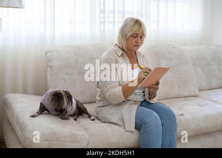 Belle femme âgée assise sur le canapé dans le salon et écrivant dans le planificateur. Banque D'Images
