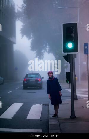 Une femme sous un manteau se tient près d'un feu de circulation dans le brouillard. Banque D'Images