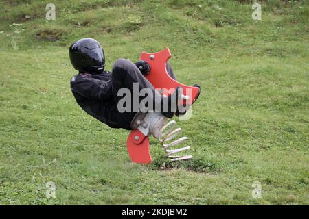 le motocycliste vêt de vêtements noirs de moto est assis sur un cheval à bascule sur l'aire de jeux pour enfants et va fou Banque D'Images