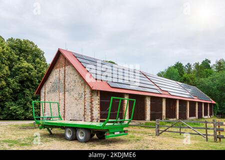 Remorque de ferme verte garée devant une grande grange. Panneaux solaires installés sur le toit de la grange Banque D'Images
