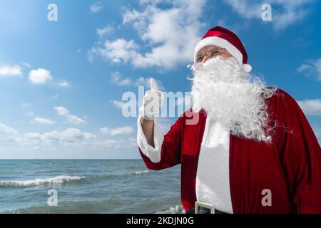 Portrait du Père Noël avec son doigt levé sur la mer un jour ensoleillé, copier l'espace. Concept de vacances de Noël ou de nouvel an dans les pays chauds Banque D'Images