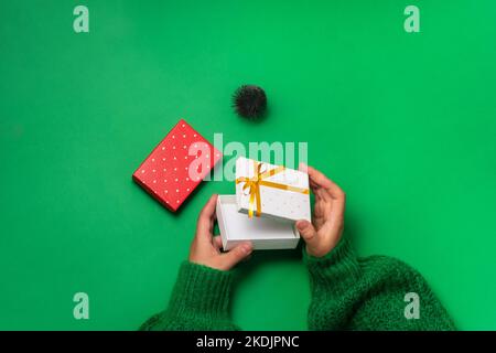 Les mains des femmes ouvrent une petite boîte cadeau blanche avec un ruban doré sur fond vert. Vue de dessus, plat, espace de copie Banque D'Images