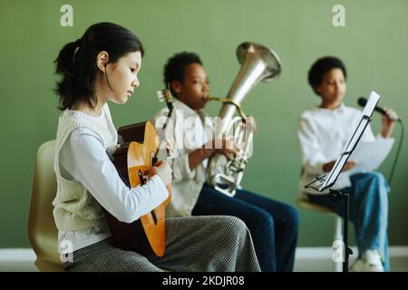 Jolie écolière asiatique jouant de la guitare acoustique et regardant des notes sur le stand de musique tout en étant assise contre deux camarades de classe en cours Banque D'Images