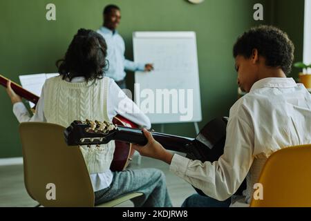 Vue arrière de deux jeunes apprenants de l'école de musique jouant des instruments à cordes devant l'enseignant debout par tableau blanc Banque D'Images