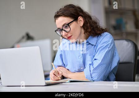 Jeune femme d'affaires regardant le webinaire en ligne au travail écrivant des notes. Banque D'Images