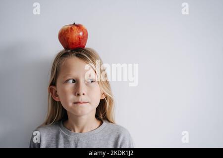 Un garçon qui se glissera sur les côtés avec une pomme sur sa tête. Sur un mur blanc. Il a de longs cheveux blonds. Banque D'Images