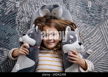 Adorable fille couchée sur le sol entourée de huskies en peluche. Sourire à l'appareil photo. Banque D'Images