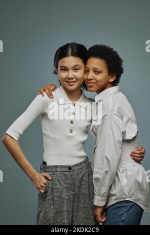 Deux adorables écolières interculturelles en blanc de style décontracté se embrassant l'une l'autre et regardant la caméra tout en posant sur fond gris Banque D'Images