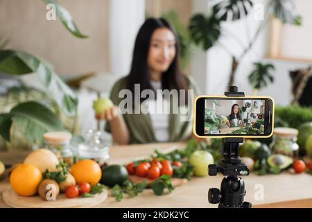 Souriante jeune femme asiatique cuisant vlogger en direct streaming son blog vidéo à des suiveurs, tout en montrant comment préparer le smoothie, la salade de pomme et d'autres ingrédients assis à table en bois Banque D'Images