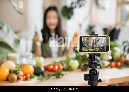 Femme asiatique faisant des aliments sains en utilisant le téléphone créant du contenu vidéo pour les médias sociaux dans le studio de cuisine de centre de villégiature tropical. Belle femme utilisant des légumes et des fruits préparant des smoothies Banque D'Images