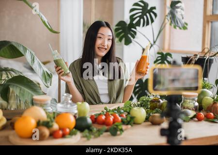 Femme asiatique faisant des aliments sains en utilisant le téléphone créant du contenu vidéo pour les médias sociaux dans le studio de cuisine de centre de villégiature tropical. Belle femme utilisant des légumes et des fruits préparant des smoothies Banque D'Images