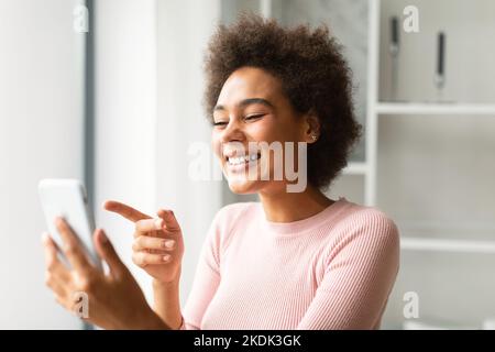Joyeuse jeune femme afro-américaine millénaire discutant au téléphone, a l'appel vidéo et le doigt de pointage à la webcam Banque D'Images