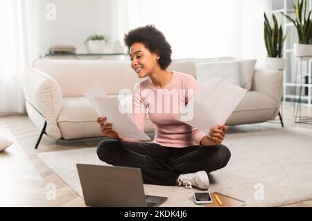 Joyeuse femme afro-américaine du millénaire occupée travaillant avec des documents, asseyez-vous au sol avec un ordinateur portable dans le salon Banque D'Images