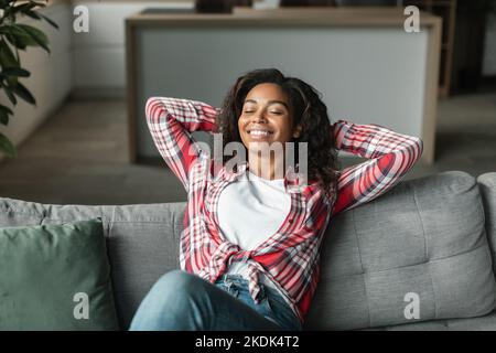 Jolie jeune femme noire souriante avec des yeux fermés, se reposer seul sur un canapé dans un intérieur minimaliste de salon Banque D'Images