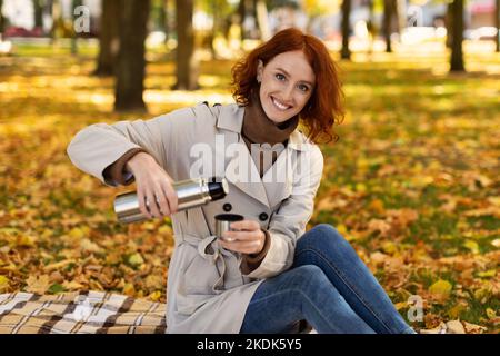 Souriante, jolie femme millénaire caucasienne avec cheveux rouges en imperméable, se trouve sur un plat, verse une boisson chaude de thermos Banque D'Images