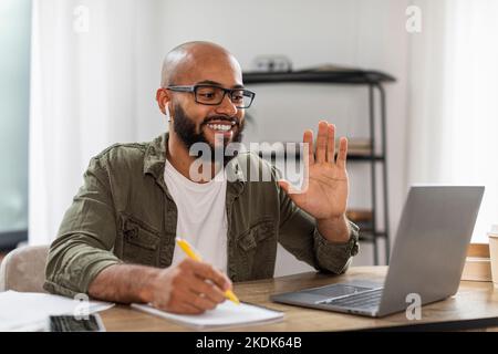 Téléconférence et télétravail. Joyeux latin assis à un bureau, ayant un appel vidéo sur un ordinateur portable, en agitant à la webcam Banque D'Images