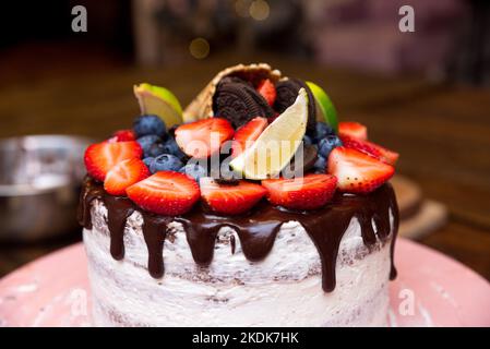 Baies fraîches sur une garniture au chocolat sur un gâteau fait maison. Gâteau d'anniversaire sur un support à gâteau rose. Avec des tranches de lime et des feuilles de menthe, des biscuits bruns et de la poudre Banque D'Images
