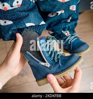 La mère met des chaussures bleues sur le pied de bébé enfant assis dans le couloir de la maison. Femme maman vêtue chaud bottes vêtements sur enfant pour l'hiver marcher dans c Banque D'Images