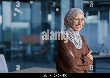 Portrait d'une femme d'affaires prospère et heureuse dans le hijab, employé de bureau souriant et regardant la caméra avec les bras croisés, travaillant dans le bureau moderne. Banque D'Images