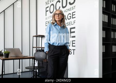 Femme d'affaires cadre dans une chemise bleue se tient au milieu du bureau et regarde la distance, caucasienne femme d'affaires d'âge moyen à Banque D'Images