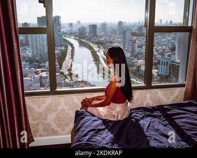 Vue latérale d'une femme asiatique assise sur un lit à la fenêtre avec vue sur Ho Chi Minh, Vietnam Banque D'Images