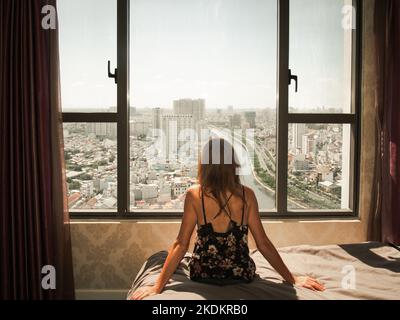 Vue arrière d'une femme caucasienne assise sur un lit et regardant à travers la fenêtre de l'horizon de Ho Chi Minh ville, Vietnam - concept d'isolement à la maison Banque D'Images