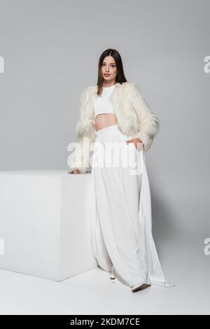 femme tendance dans une veste en fausse fourrure tendance et tenue blanche totale debout avec main dans la poche près du cube sur le gris Banque D'Images