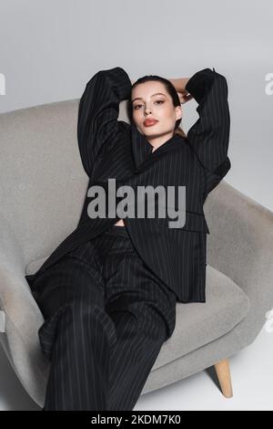 Jeune femme tendance en costume rayé noir regardant l'appareil photo sur un fauteuil sur fond gris Banque D'Images