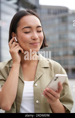 Prise de vue verticale d'une femme asiatique souriante dans un casque sans fil, aime écouter de la musique dans un casque, tient le téléphone portable Banque D'Images