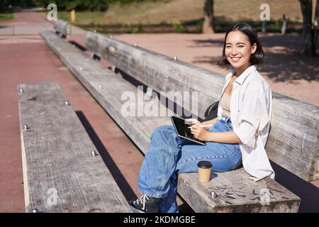 Portrait d'une fille asiatique assis sur un banc dans le parc, dessiner sur sa tablette numérique avec un stylo, sourire heureux, devenir créatif Banque D'Images