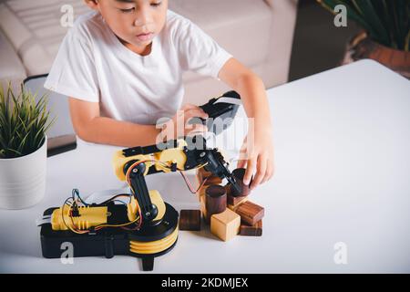 Joyeux petit garçon asiatique utilisant la télécommande jouant le bras de machine robotique pour ramasser le bloc de bois Banque D'Images