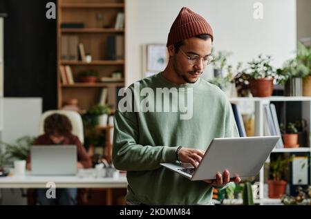 Jeune responsable d'assistance informatique sérieux avec ordinateur portable répondant aux questions des utilisateurs en ligne en se tenant devant l'appareil photo dans un espace de travail Banque D'Images