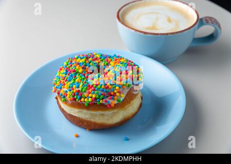 Beignet couvert de sucre glace et de saupoudrés multicolores sur une assiette bleue et une tasse de cappuccino avec mousse de lait sur une table blanche, vue normale. Copier l'espace Banque D'Images
