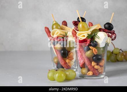 Jarcuterie en deux verres comprend différents types de saucisses, de cornichons, de fromage, de noix, de légumes et de fruits. Version moderne d'en-cas festifs sur fond gris Banque D'Images