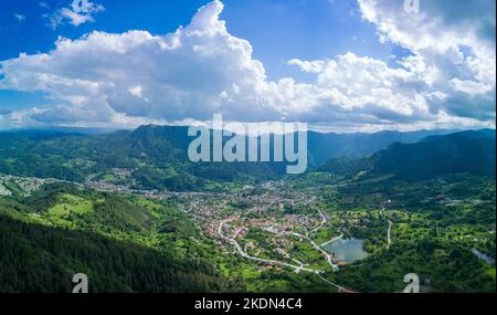 Petite ville bulgare Smolyan avec maisons et lac est couverte de végétation et de rayons de soleil sous les nuages. Montagnes Rhodope. Panorama, vue de dessus Banque D'Images