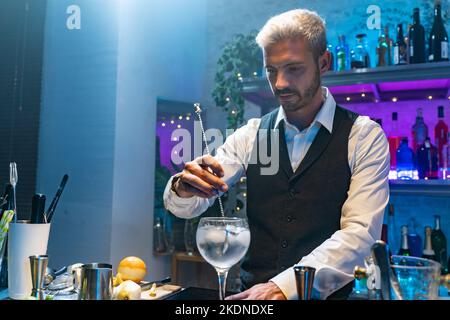 Un jeune barman dans une chemise blanche et un tablier noir fait un cocktail à la fête dans la boîte de nuit avec des lumières colorées. Concept de vie nocturne. Banque D'Images