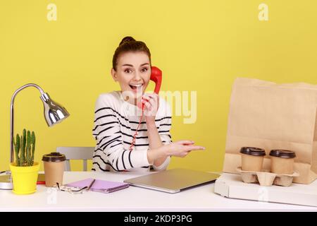 Bonne femme pointant vers une boîte à pizza tasses de café et sac de papier avec achat avec sourire et téléphone fixe parlant, appréciant la livraison rapide de nourriture. Studio d'intérieur tourné isolé sur fond jaune Banque D'Images