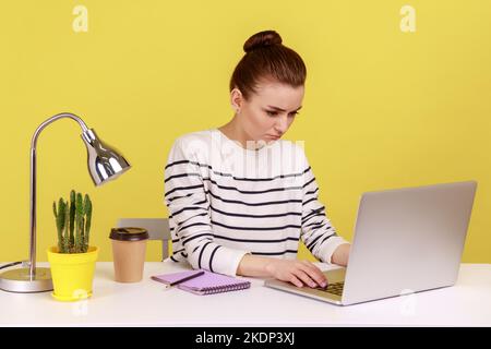 Triste femme malheureuse gestionnaire de bureau assis sur le lieu de travail regardant l'écran d'ordinateur portable ayant des appels vidéo, tapant sur le clavier avec l'expression contrariée. Studio d'intérieur tourné isolé sur fond jaune. Banque D'Images