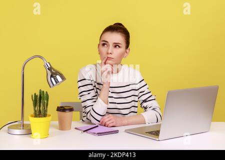 Femme pensive en chemise à rayures, assise avec une expression réfléchie et confuse tout en travaillant sur un ordinateur portable au bureau, pensant à un nouveau projet. Studio d'intérieur tourné isolé sur fond jaune. Banque D'Images