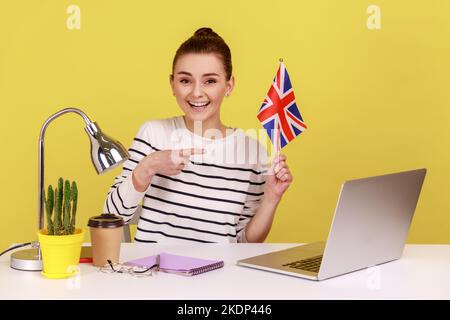 Souriante femme positive assise sur le lieu de travail avec, pointant sur le drapeau du Royaume-Uni de Grande-Bretagne, exprimant le bonheur, relocalisation à l'étranger. Studio d'intérieur tourné isolé sur fond jaune. Banque D'Images